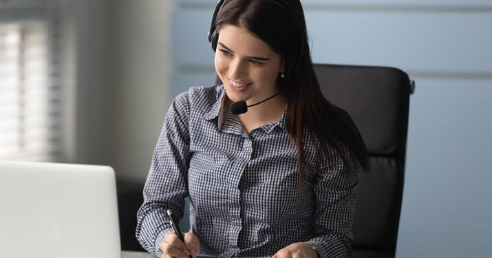 Smiling businesswoman watching educational webinar, writing down notes. Happy young female professional employee worker wearing headset with microphone, holding video call with partners or clients.